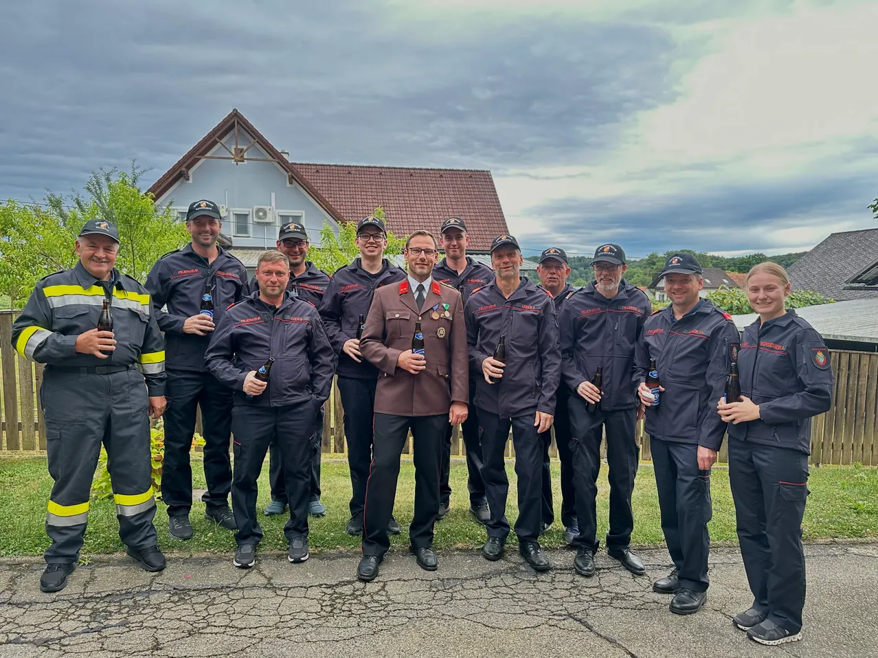 Eine Gruppe Feuerwehrleute in Uniform steht auf der Straße und hält Bierflaschen in der Hand, mit einem Haus im Hintergrund unter einem bewölkten Himmel.