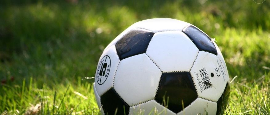 Ein weißer und schwarzer Fußball mit einem runden Logo und Barcode liegt auf einem Grasfeld unter Sonnenlicht.