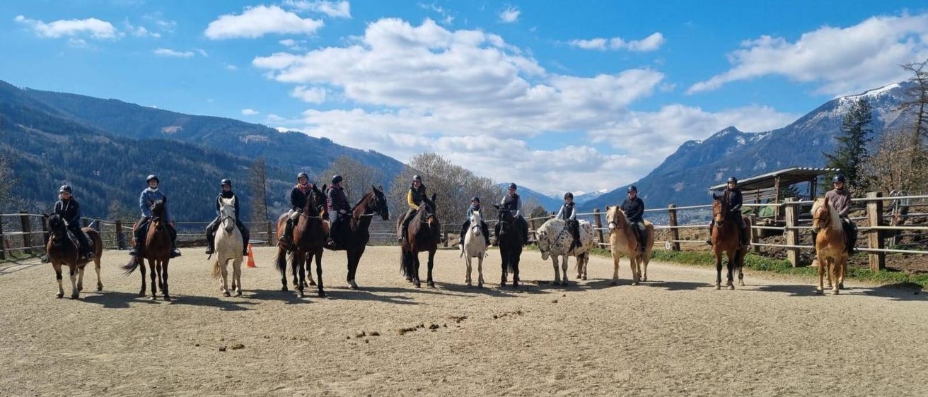 Eine Gruppe von Menschen reitet Pferde in einem Außenbereich, Berge im Hintergrund und ein Zaun sichtbar.