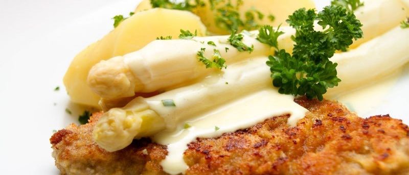 Nahaufnahme eines Gerichts aus paniertem Kalbfleisch mit weißer Sauce, serviert mit Spargel und Kartoffeln.