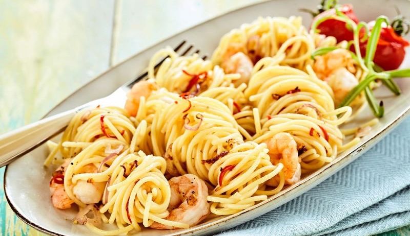 Ein Teller mit Spaghetti und Garnelen, garniert mit Chili, Tomaten und einer Gabel, auf einem blauen Tuch auf einem Holztisch.