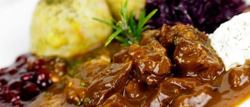 Ein weißer Teller hat ein Fleischgericht mit Soße, ein Stück Brot und etwas Rotkohl.