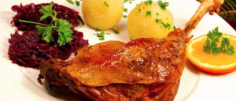 Ein Teller mit knusprigem gebratenem Entenschenkel, serviert mit zwei gelben Kartoffelklößen, garniert mit Petersilie, und einer Beilage aus lila Sauerkraut. Eine Orangenscheibe befindet sich auf der rechten Seite.