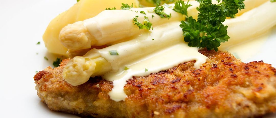 Nahaufnahme eines Tellers mit golden gebräuntem, paniertem Kalbsschnitzel, überzogen mit cremiger weißer Sauce und Spargel. Garniert mit Petersilie.