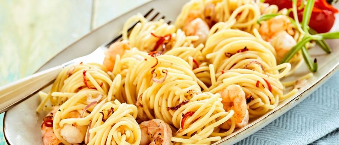 Ein Teller mit Pasta, Garnelen, roten Chiliflocken und Zwiebeln auf einem blauen Tuch mit einer Gabel.