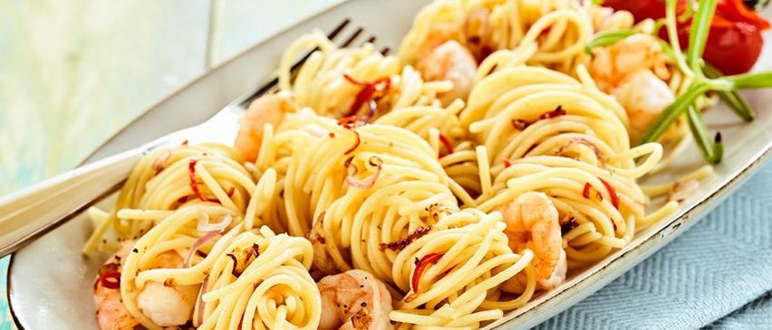 A plate of pasta with shrimp, red chili flakes, and onions on a blue cloth with a fork.