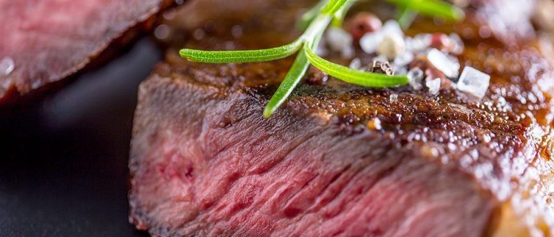 Eine Nahaufnahme eines Steaks mit einem Rosmarinblatt und Gewürzen. Das Steak ist an den Rändern leicht angebrannt.