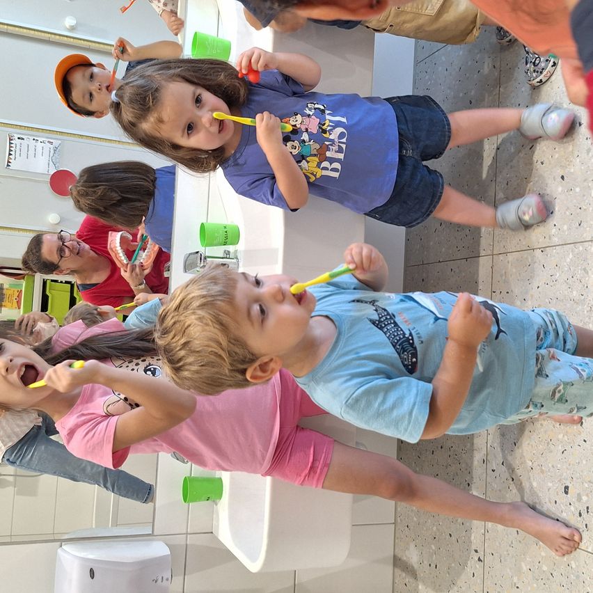 Mehrere Kinder, mit Zahnbürsten in der Hand, putzen ihre Zähne in einem Badezimmer. Hinter ihnen beaufsichtigt ein Mann. Die Kinder schauen in die Kamera und lächeln.