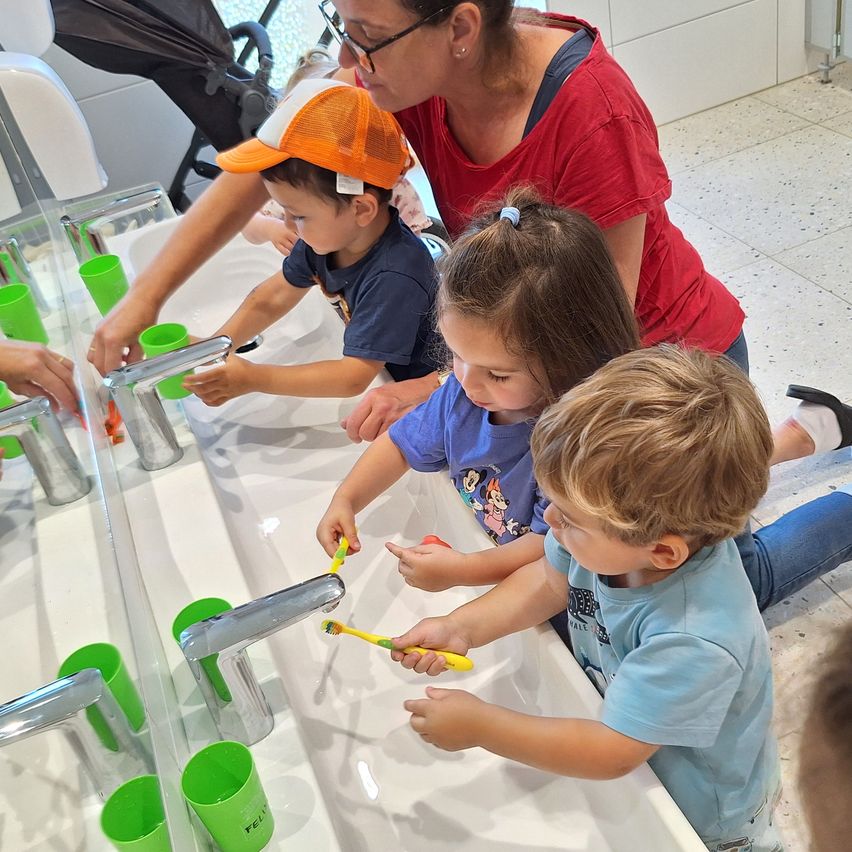 Eine Frau und drei Kinder waschen sich die Hände an einem Waschbecken, mit grünen Bechern und gelben Zahnbürsten. Die Kinder sitzen auf dem Boden.