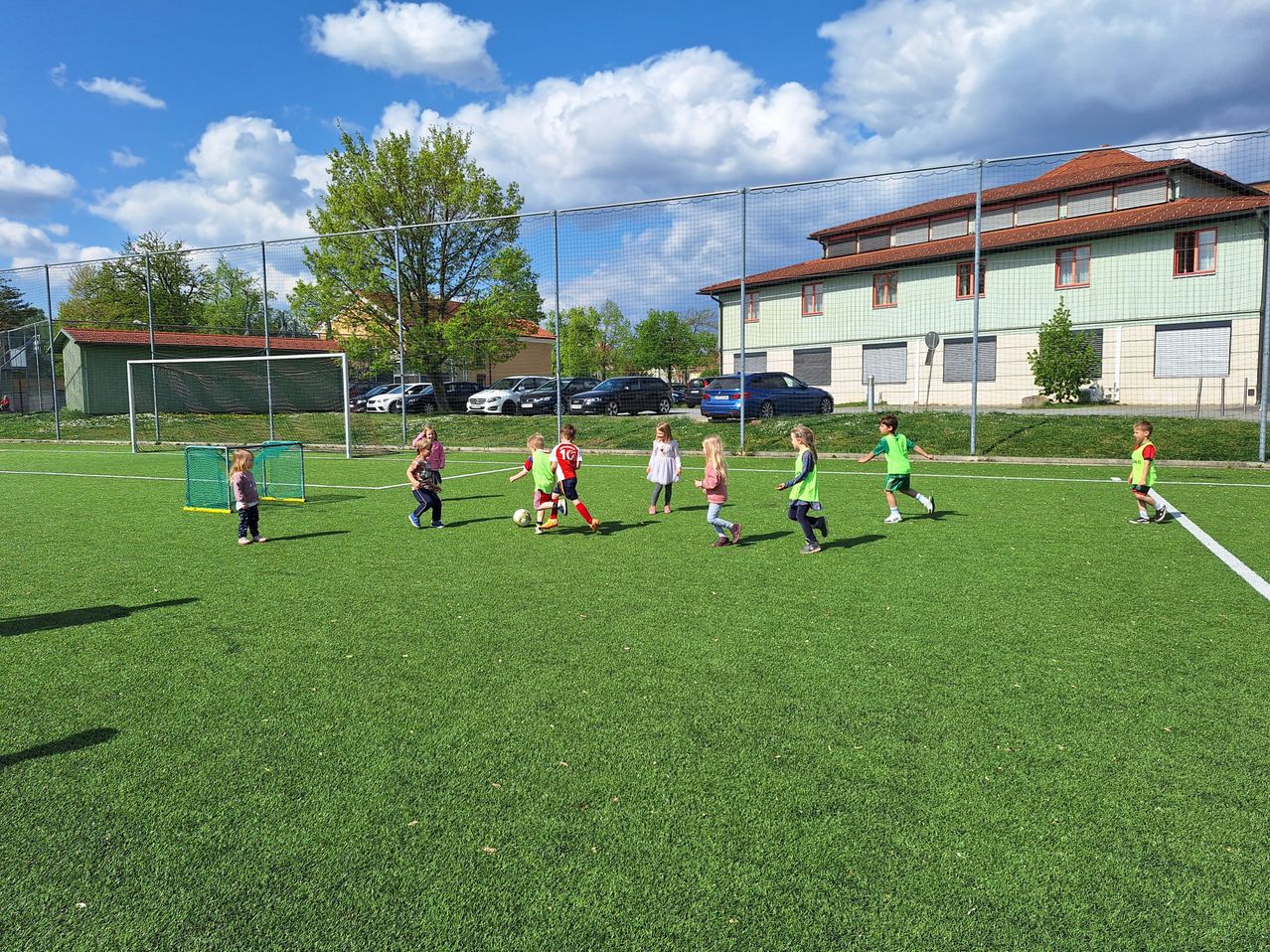 Kinder spielen Fußball auf einem Feld mit grünem Kunstrasen. Zwei Teams in grünen und roten Trikots. Ein Gebäude und ein Zaun im Hintergrund.