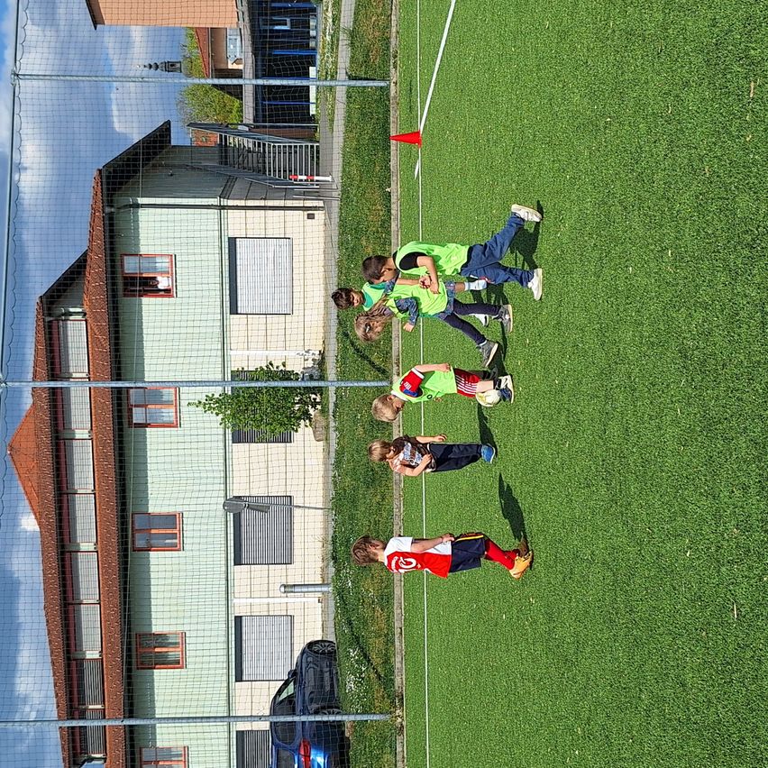 Mehrere Kinder in grünen Trikots spielen Fußball auf einem Feld, mit einem Haus und einem geparkten Auto in der Nähe.