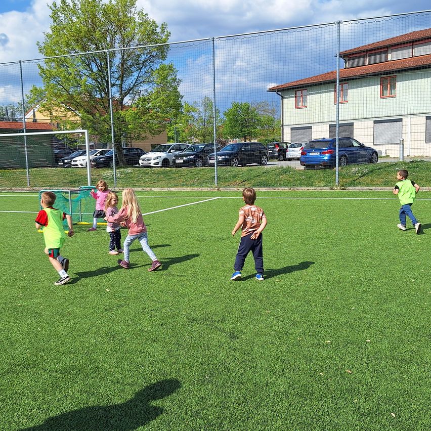 Kinder spielen auf einem Fußballfeld. Ein Kind läuft auf das Tor zu. Hinter ihnen steht ein Zaun mit Netz. Mehrere Autos sind auf dem Parkplatz geparkt.