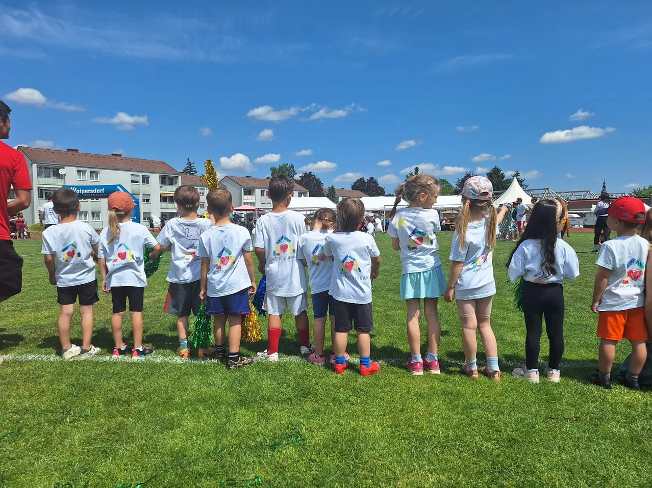 Eine Gruppe junger Kinder in passenden T-Shirts steht auf einem Rasenfeld, wahrscheinlich warten sie auf den Start eines Rennens. Einige tragen Mützen, und einige haben Pompons. Im Hintergrund befinden sich Gebäude und Bäume.