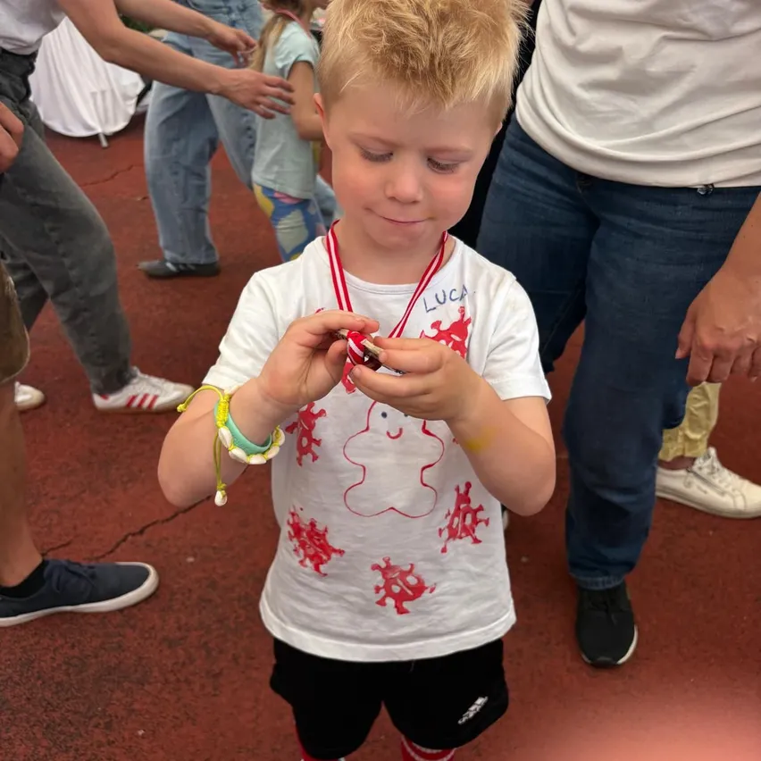 Ein junger Junge mit blonden Haaren hält ein kleines rotes Objekt in den Händen und trägt eine Medaille. Er ist von Menschen bei einer Veranstaltung umgeben.