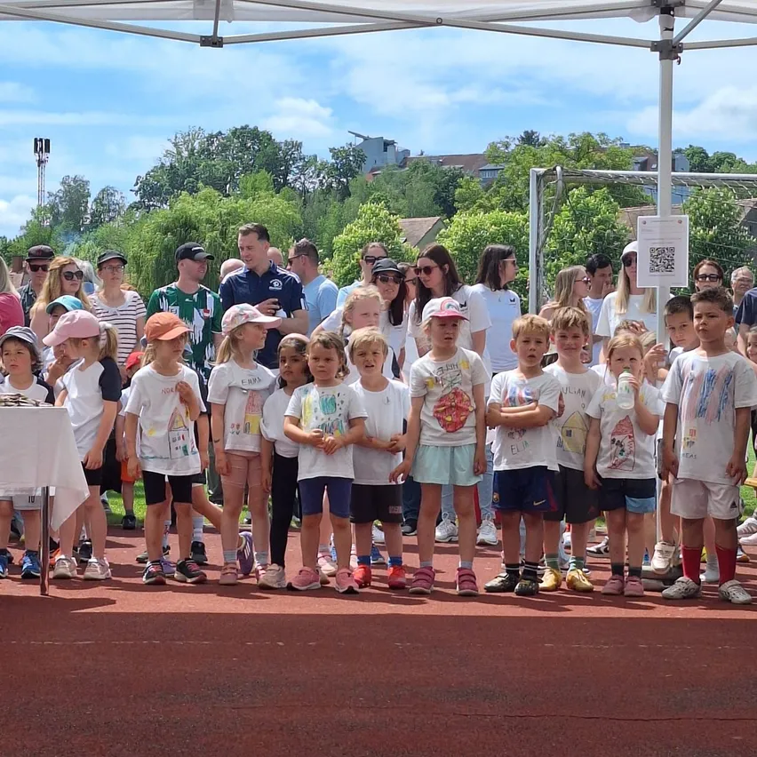 Kinder in weißen Shirts stehen auf einer Laufbahn unter einem Schirm, mit Erwachsenen und Bäumen im Hintergrund. Einige tragen Hüte und Turnschuhe.