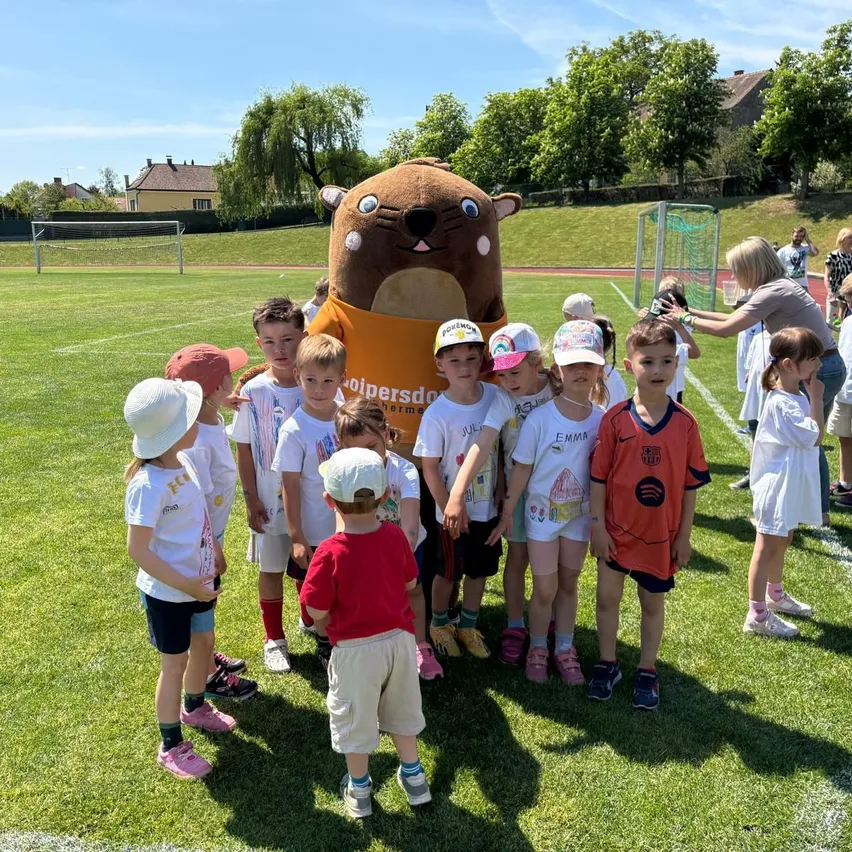 Kinder in Sportkleidung versammeln sich um eine Maskottchen auf einem sonnigen Feld, eine Frau mit einem Becher in der Nähe.