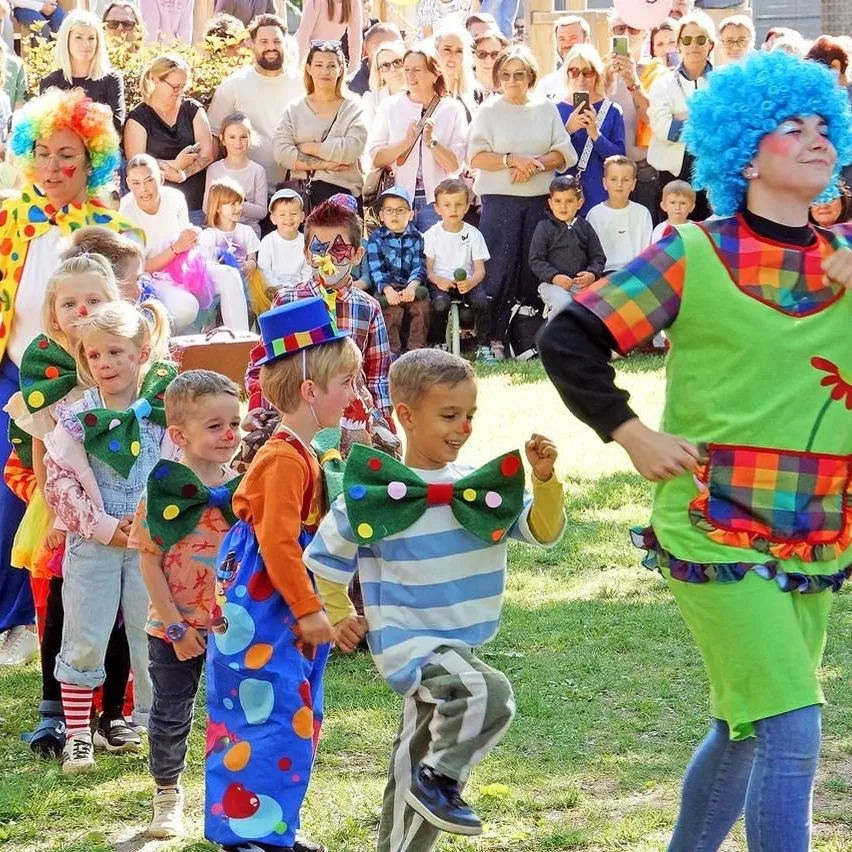 Mehrere Kinder in Clownkostümen gehen mit einem Clown auf dem Gras. Ein Publikum aus Erwachsenen und Kindern sieht ihnen zu.