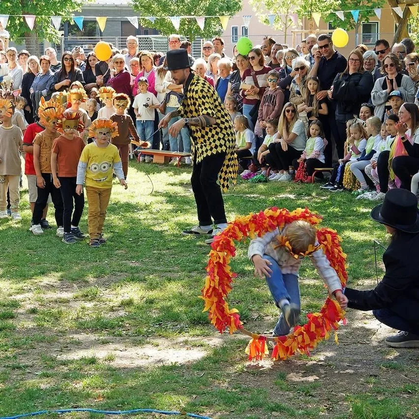 Eine Menschenmenge, darunter Kinder mit Tiermasken, beobachtet einen Mann, der einen Jonglierakt mit einem großen orangefarbenen Hula-Hoop vorführt. Die Kinder stehen in einer Reihe, und der Mann kniet sich hin, um einem Kind beim Durchgehen des Reifens zu helfen.
