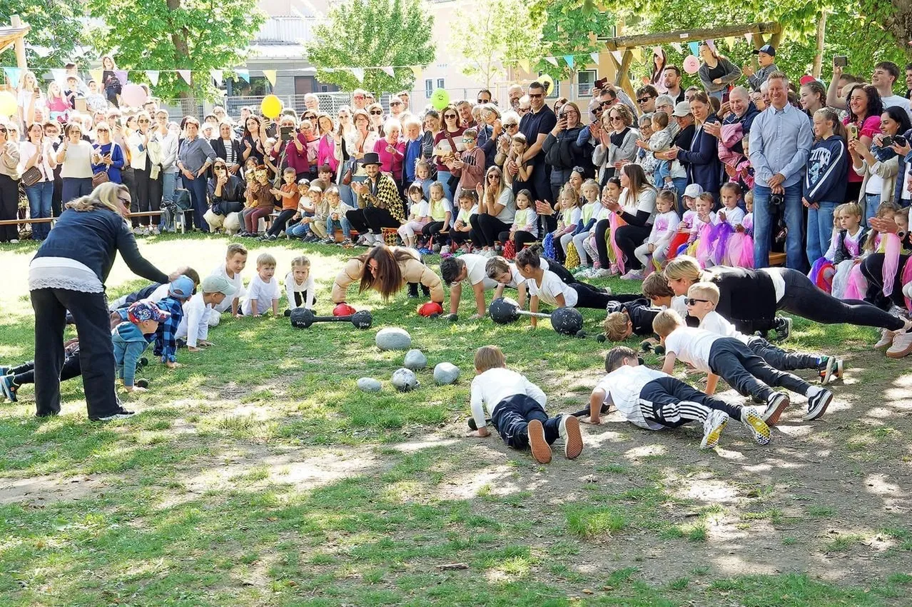 Eine große Menschenmenge, darunter auch Kinder, versammelt sich im Freien. Einige von ihnen machen Übungen mit Bällen auf dem Rasen. Einige Leute stehen hinter ihnen, beobachten und klatschen.