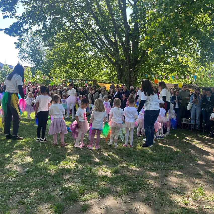 Eine Gruppe von Kindern in weißen T-Shirts und bunten Tüllröcken führt in einem Park vor, umgeben von Zuschauern unter einem großen Baum.