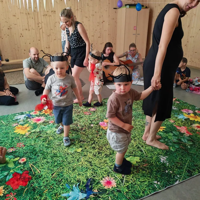 Eine Gruppe von Kindern und Erwachsenen versammelt sich in einem Raum mit einem Blumenteppich. Zwei Kinder mit Kopfbändern halten sich an den Händen von Erwachsenen, während andere sitzen oder im Hintergrund spielen.