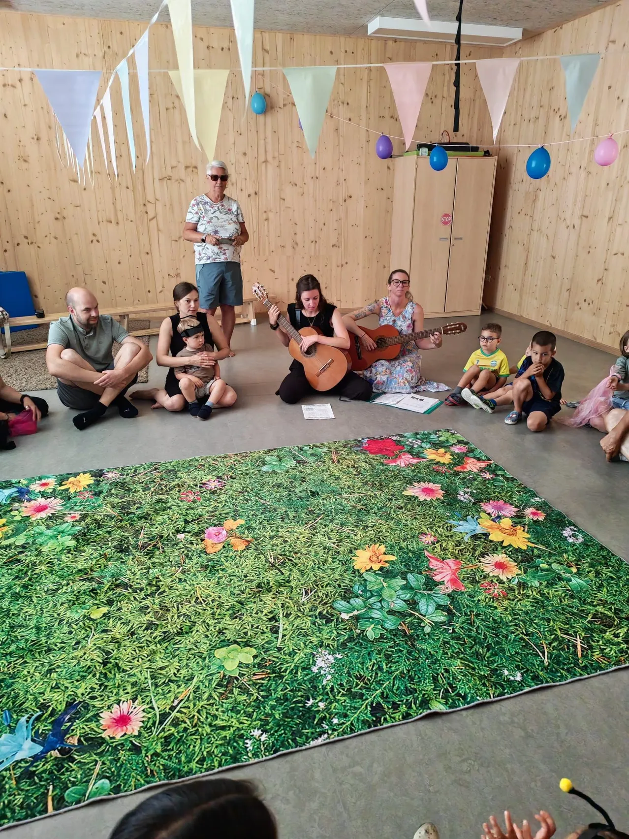 Eine Gruppe von Menschen sitzt auf dem Boden in einem Raum mit Holzwänden. Zwei Frauen spielen Gitarre. Ein farbenfroher Blumenteppich liegt auf dem Boden.