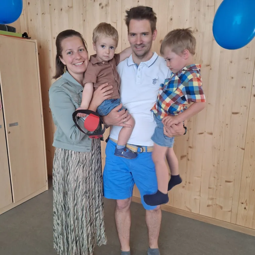 Ein Familienfoto mit zwei jungen Jungen und ihren Eltern. Der Vater hält einen Jungen, während die Mutter den anderen hält. Sie stehen vor einer Holzwand mit Luftballons.