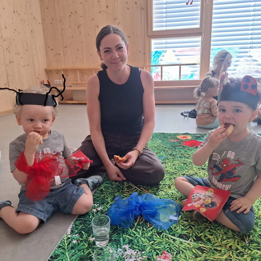 Eine Frau und zwei Kinder sitzen auf einer Matte und essen. Der Junge trägt eine Spinnenkopfbedeckung. Die Frau trägt ein schwarzes Tanktop und sitzt kreuzbeinisch.
