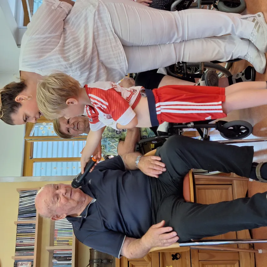 Ein junger Junge in einem roten und weißen Fußballtrikot hält ein Mikrofon, während er auf dem Schoß eines Mannes sitzt, der lächelt und die Hand des Jungen hält. Eine Frau in einem weißen Shirt beugt sich über ihn. Dahinter steht ein Regal mit vielen Büchern.
