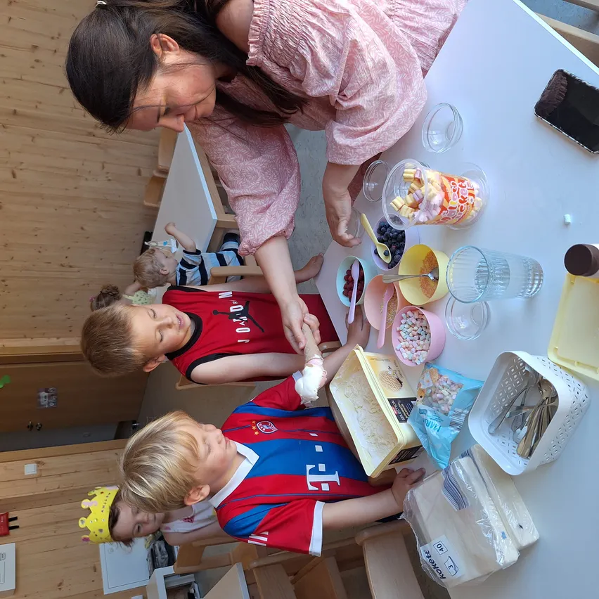 Eine Frau hilft zwei Jungen beim Zubereiten von Essen an einem Tisch. Schüsseln, Löffel und Süßigkeiten sind auf dem Tisch. Die Jungen tragen Sporttrikots. Ein weiteres Kind sitzt am anderen Tisch im Hintergrund.