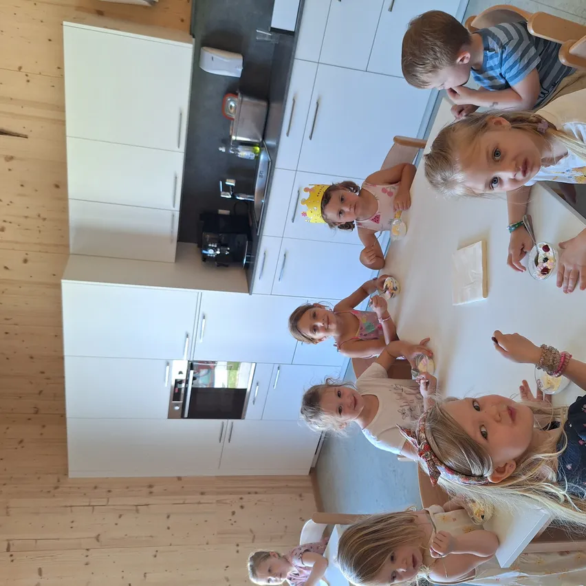 Mehrere kleine Kinder sitzen um einen Tisch in einer Küche und lächeln und essen. Sie sind von weißen Schränken und einem Holzboden umgeben.