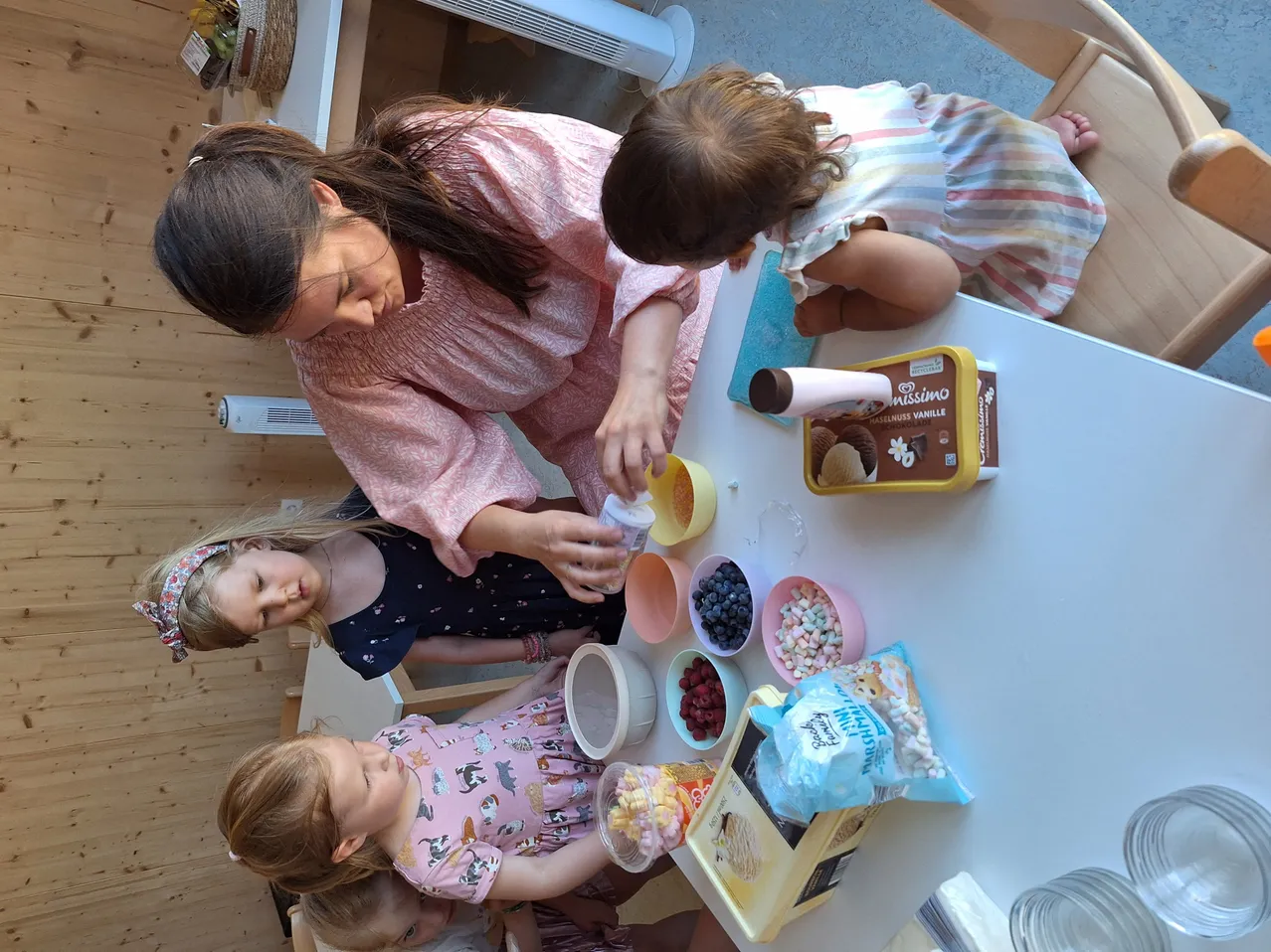 Eine Frau und drei Mädchen sitzen um einen Tisch herum. Die Frau gießt eine Substanz in eine Schüssel, und die Mädchen beobachten. Verschiedene Lebensmittel und Utensilien sind auf dem Tisch.
