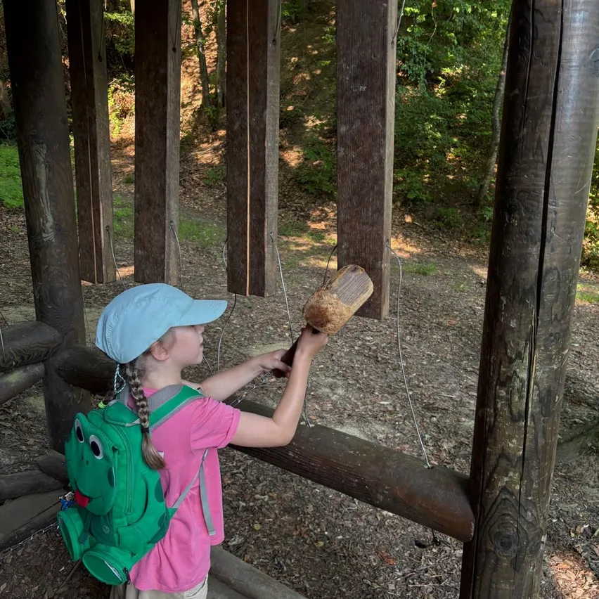 Ein kleines Mädchen in einem rosa Shirt und Hut schlägt mit einem Hammer eine Holzglocke. Sie trägt einen grünen Rucksack. Der Ort ist draußen mit Holzpfosten.