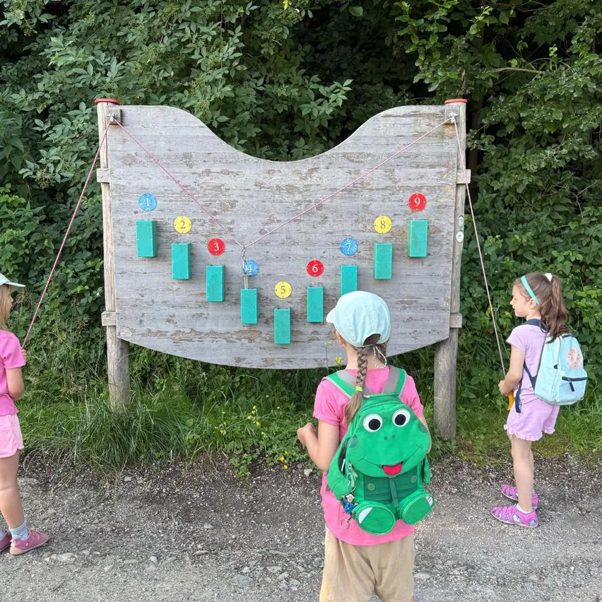 Drei Kinder stehen neben einem Holzspielbrett mit den Nummern 1-9 in einem Wald. Ein Mädchen trägt einen Froschrucksack.