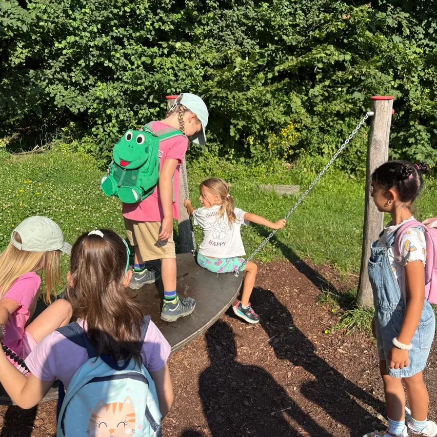 Kinder spielen auf einem Wippschaukel in einem Grasbereich, eine Mädchen auf der Schaukel und andere beobachten. Ein Kind trägt einen Rucksack mit Froschdesign.