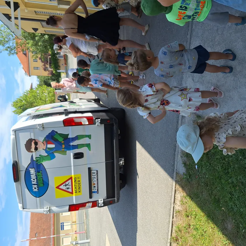 Eine Gruppe von Kindern und Erwachsenen versammelt sich um einen Van, wobei mehrere Personen Mützen und Turnschuhe tragen. Der Van hat einen Cartoon-Charakter auf der hinteren Tür, ein Nummernschild und Warnschilder.
