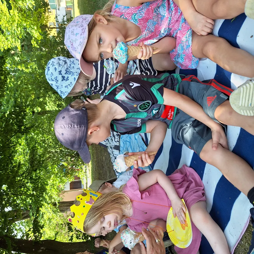 Eine Gruppe von Kindern liegt draußen auf einer Decke und genießt Eis unter einem Baum. Ein Kind trägt eine Krone und hält einen Papierteller.