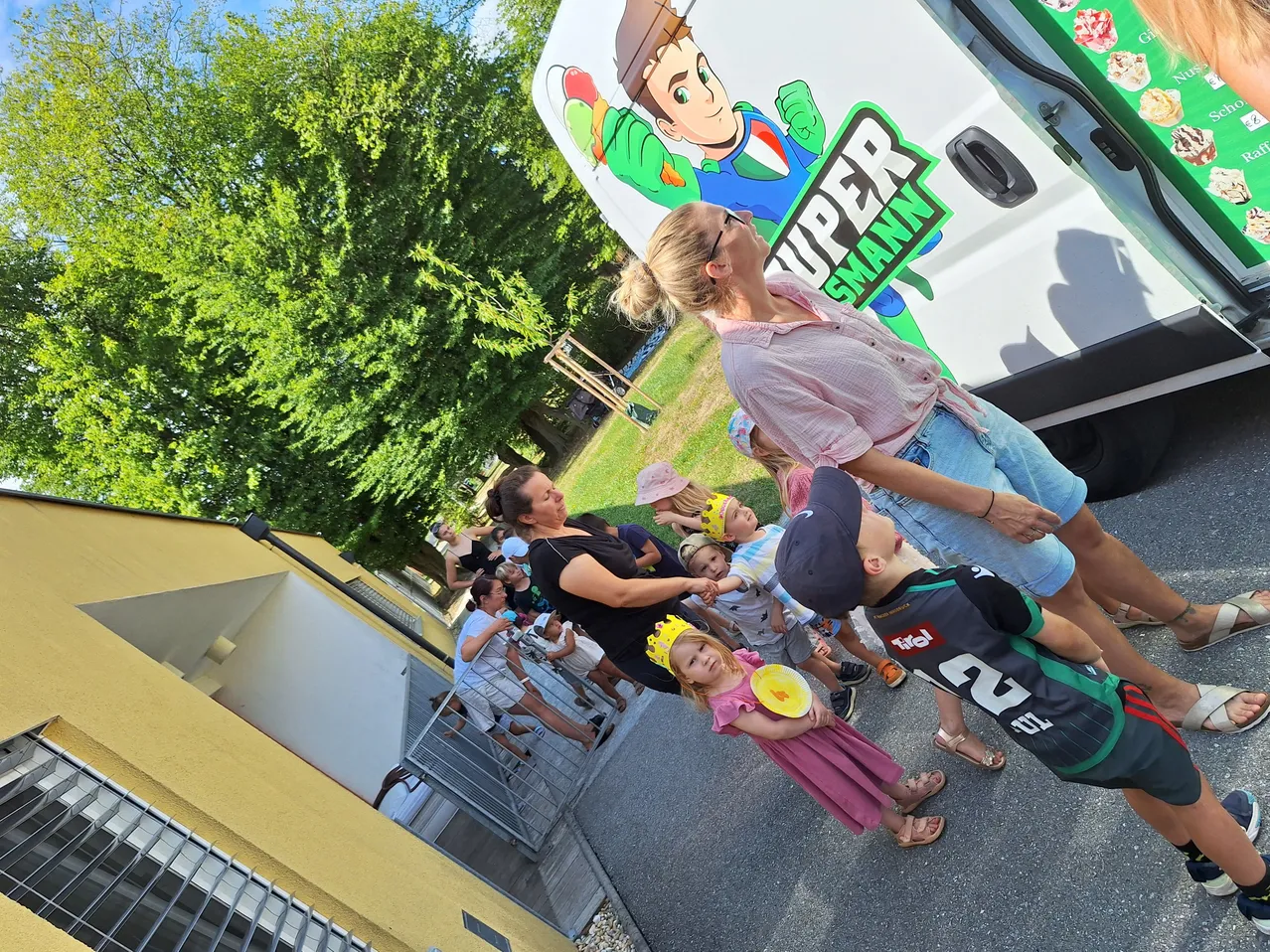 Eine Gruppe von Kindern und Erwachsenen, einige mit Kronen, stehen vor einem Van mit einem Comic-Charakter. Bäume und ein Gebäude sind im Hintergrund zu sehen.