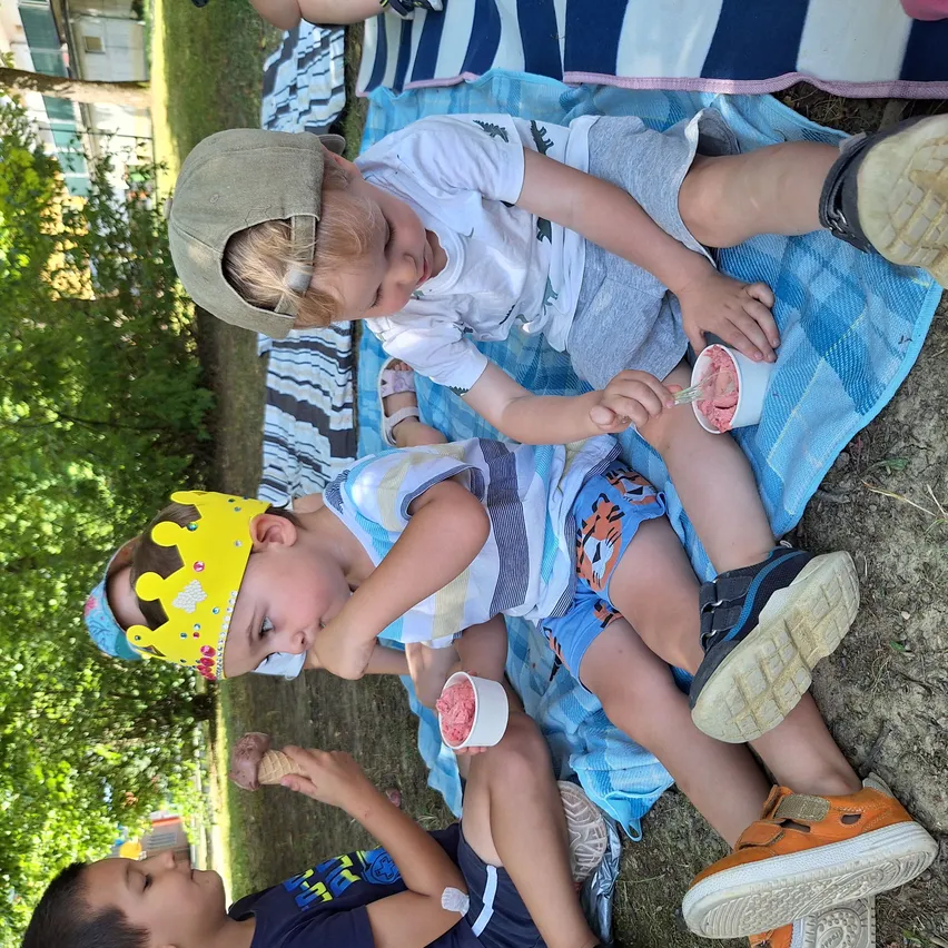 Drei Kinder in lässiger Sommerkleidung sitzen auf einer Decke. Sie haben Eis und schauen zur Kamera. Eins trägt eine Mütze, eins eine Krone und ein anderes eine Gesichtsmaske.