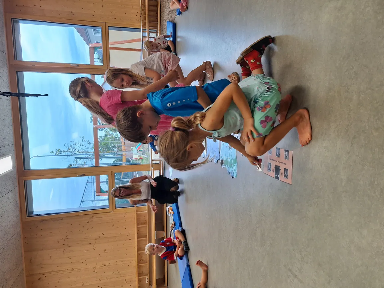 Kinder spielen auf dem Boden in einem Raum mit Glasfenstern und einer Holzwand. Eine Frau lächelt sie an.