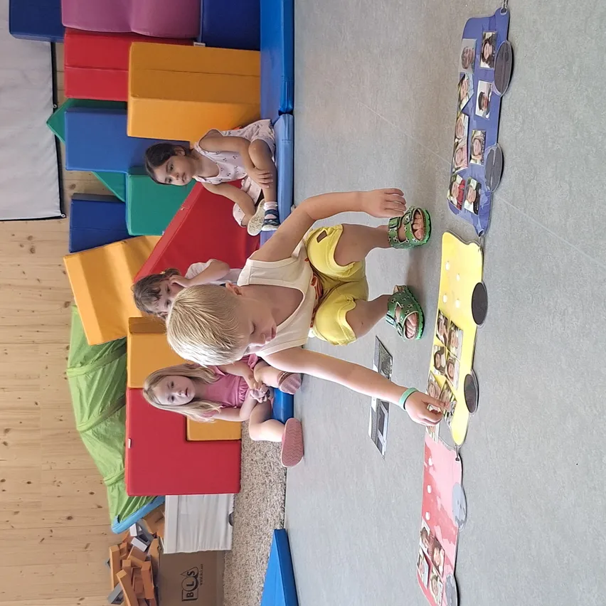 Kinder spielen in einem Raum mit bunten Bauklötzen, ein Junge lehnt sich mit einer Fototafel an eine Wand, andere spielen herum.