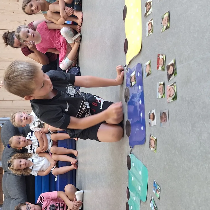 Kinder sitzen in einem Kreis um eine Wand mit einem bunten Zug. Der Junge in der Mitte trägt ein schwarzes T-Shirt. An der Wand sind Fotos und Zugteile befestigt.