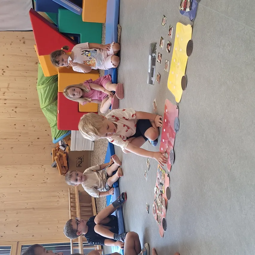 Mehrere Kinder spielen mit Spielzeug und einem Puzzle auf dem Boden. Sie schauen sich das Puzzle an, das an der Wand befestigt ist. Der Raum hat Holzwände und wird von natürlichem Licht beleuchtet.
