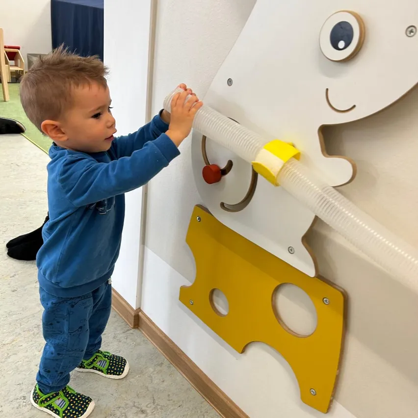 Ein kleiner Junge spielt mit einem an der Wand montierten interaktiven Spielzeug, das eine große gelbe Käseform mit Löchern und einem roten Knopf aufweist. Er hält eine durchsichtige Röhre, möglicherweise bläst er hinein.