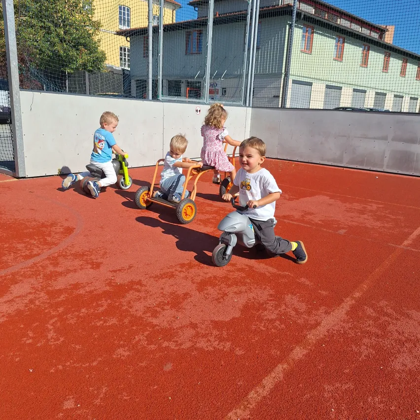 Vier Kinder fahren auf einem roten Untergrund mit Spielzeug-Dreirädern, im Hintergrund ein Gebäude und ein Zaun.