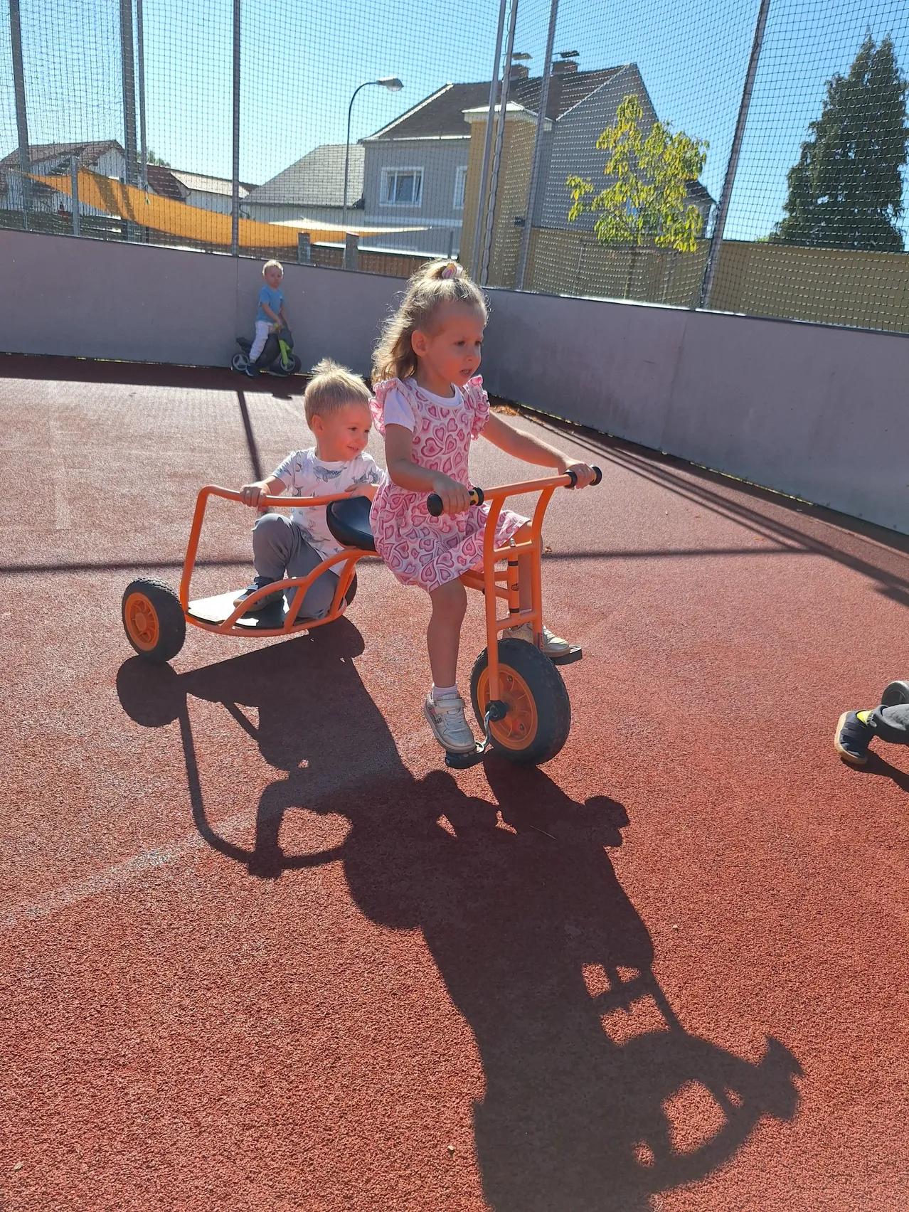 Zwei kleine Kinder fahren auf einer roten Laufbahn mit einem Dreirad, wobei ein Junge vorne und ein Mädchen dahinter sitzt. Ein weiteres Kind fährt im Hintergrund mit einem Roller.
