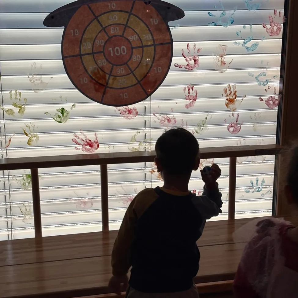 Ein junger Junge steht vor einem Fenster mit einer Hexenmütze und einem Dartboard. Das Fenster hat einen weißen Rollladen und ist mit Handabdrücken in verschiedenen Farben verziert.
