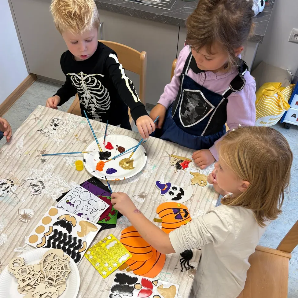 Drei Kinder sitzen um einen Tisch herum und beschäftigen sich mit einer Malaktivität. Der Tisch ist mit einem Tuch bedeckt und hat verschiedene Halloween-Themen, einschließlich Kürbisse, Spinnen und Skelette. Die Kinder halten Pinsel und scheinen auf Papiertellern zu malen.