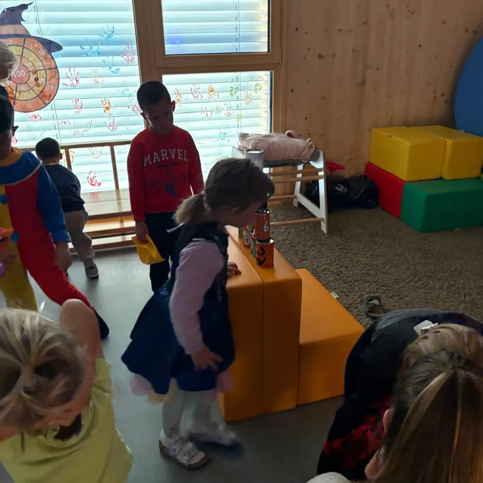 Eine Gruppe von Kindern spielt in einem Raum mit Holzwänden und einem Fenster. Ein Mädchen in einem blauen Kleid schaut auf etwas auf einem Tisch.