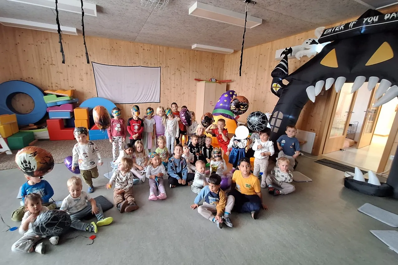 A group of children are sitting on the floor in a room with wooden walls. They are wearing costumes and holding balloons.